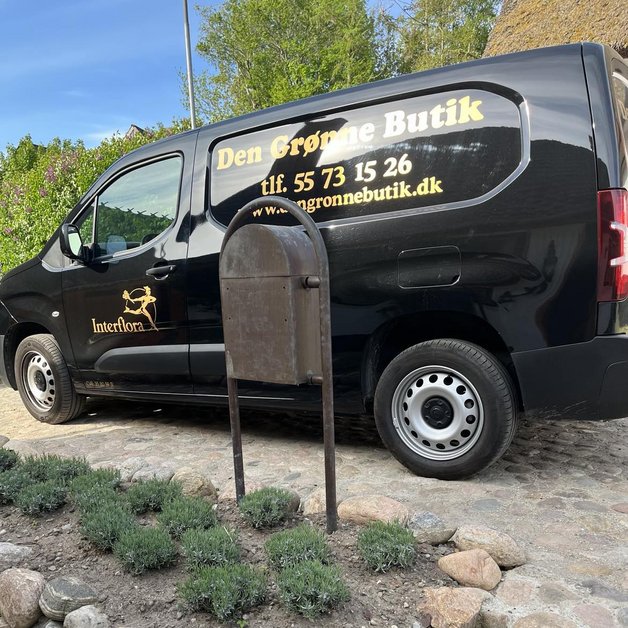 En sort varevogn med teksten "Den Grønne Butik" står parkeret ved siden af en affaldsbeholder. I forgrunden er der en blomsterbed med lavendel og store sten. Baggrunden viser grønne træer og en grusvej.