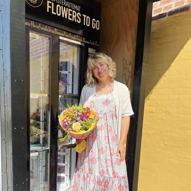 En kvinde står foran en blomsterautomat med en farverig buket blomster i hånden. Hun bærer en lys kjole med mønster og ser glad ud. Bag hende er automaten, der tilbyder blomster til salg.